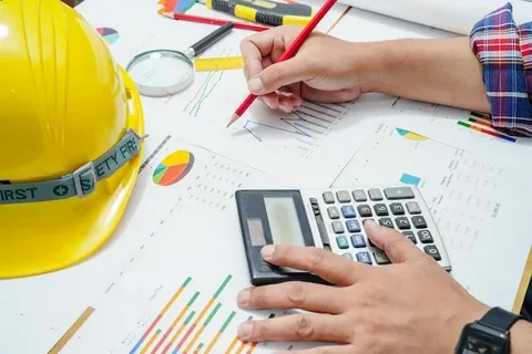 Construction hard hat, hands using a calculator and pencil over financial charts and blueprints for detailed project cost estimating.