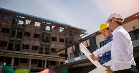 Two construction managers in hard hats reviewing blueprints with a large commercial building under construction in the background.
