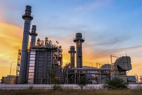 A large industrial power plant or factory with tall smokestacks and piping, set against a dramatic sunset sky.