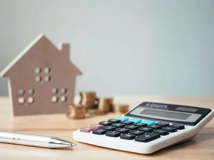 Miniature wooden house, stack of coins, calculator, and pen on a desk, representing preliminary cost estimating for residential projects.