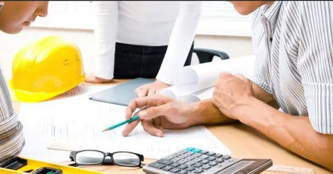 Construction professionals reviewing blueprints with a calculator, hard hat, and tools on a desk for preliminary project estimating.