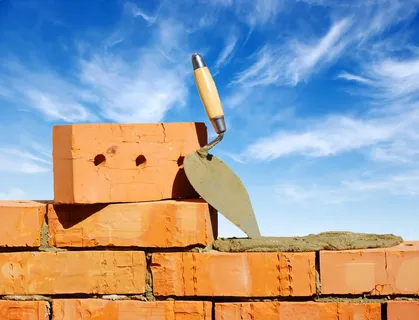 Close-up of a brick wall under construction with a trowel resting on wet mortar, set against a bright blue sky.
