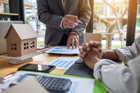 Business professionals reviewing financial charts with a miniature house model, calculator, and phone during an opening project estimating meeting.